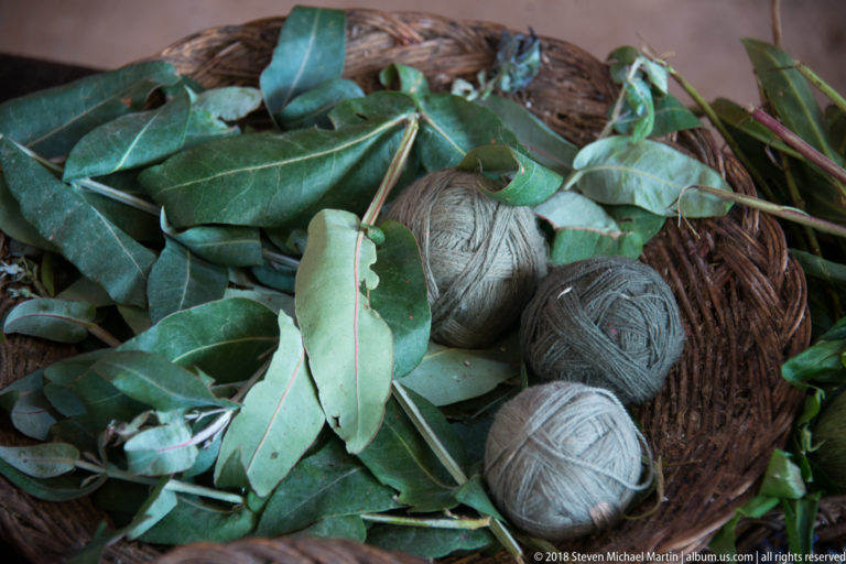 Fresh green leafy branches arranged over several balls of gray, green, and pale yarn in a woven basket.