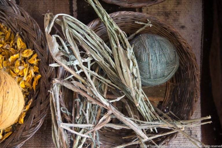 Dried stems and leaves in a woven basket beside a muted green-gray ball of yarn, with yellow plant material at left.