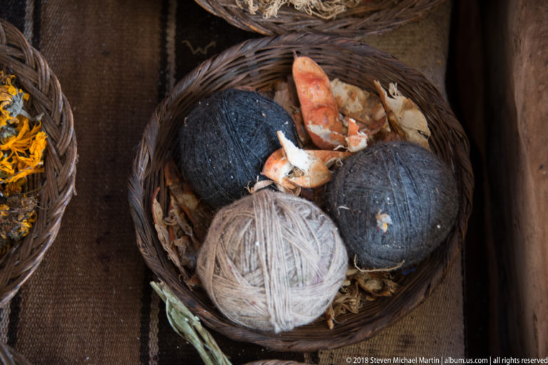 Three large balls of dark and pale gray yarn in a woven basket with dry plant fragments and orange-red material.