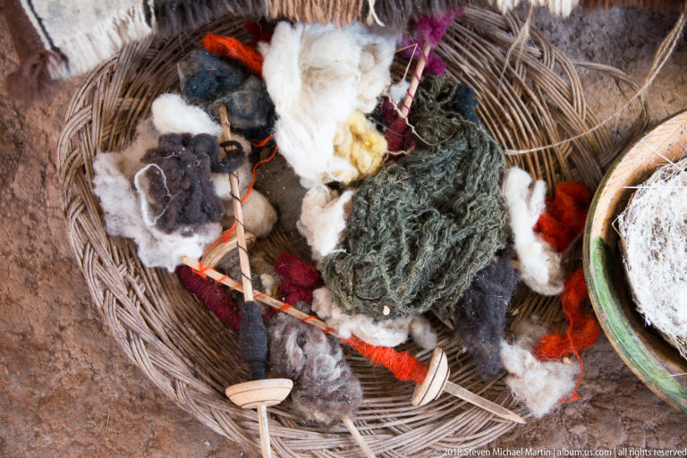Overhead view into a woven basket containing loose white, gray, green, red, and yellow fibers with small wooden tools.
