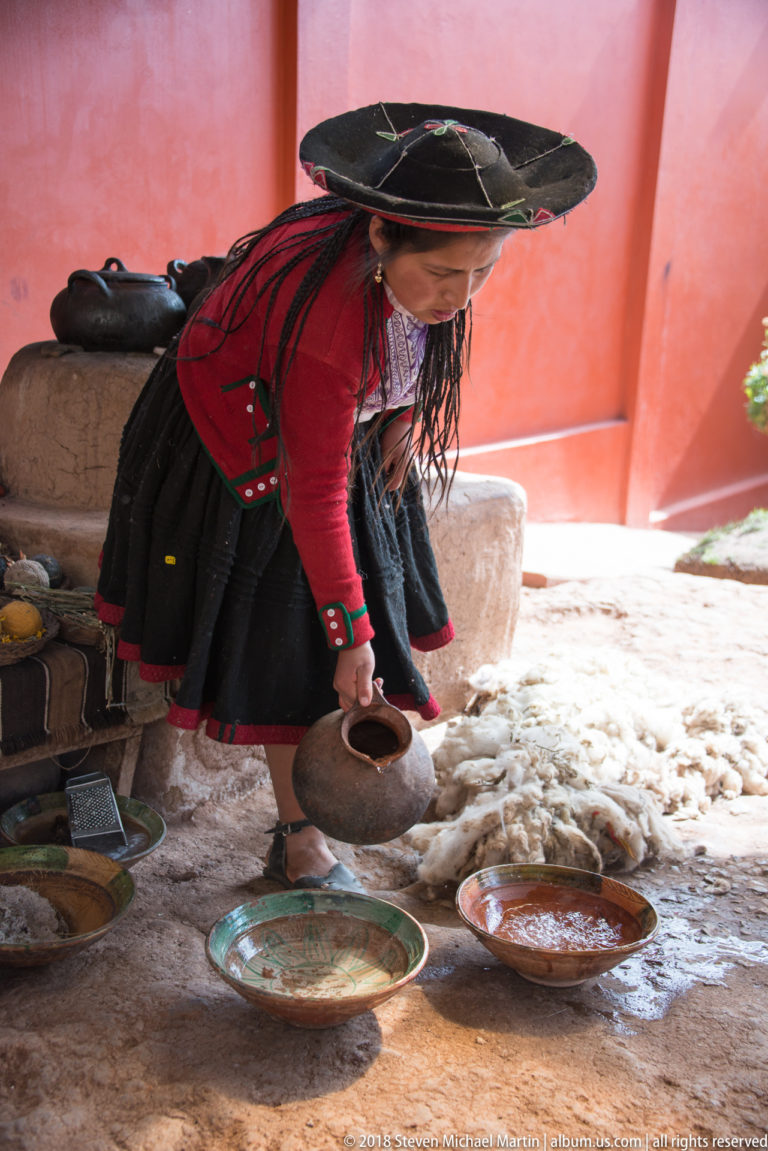 Woman in traditional highland clothing bending over shallow dishes while tipping a dark container, with wool fleece behind her.
