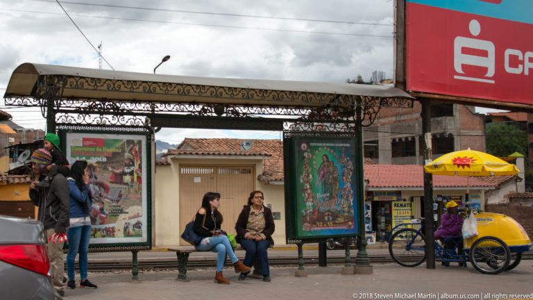 Ornate metal bus shelter with seated and standing passengers, advertising panels, and a yellow-canopied vendor cart.