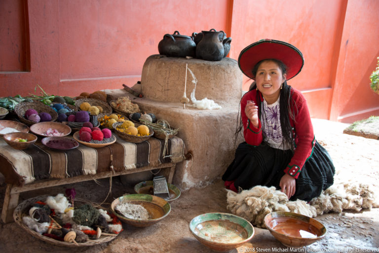 Woman in a broad red hat seated beside dyed yarn, wool fleece, bowls, dark vessels, and a small clay hearth.