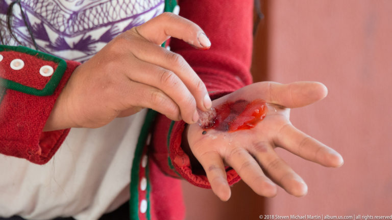 Open palm holding a small amount of vivid red crushed or moist dye material, touched by the other hand.