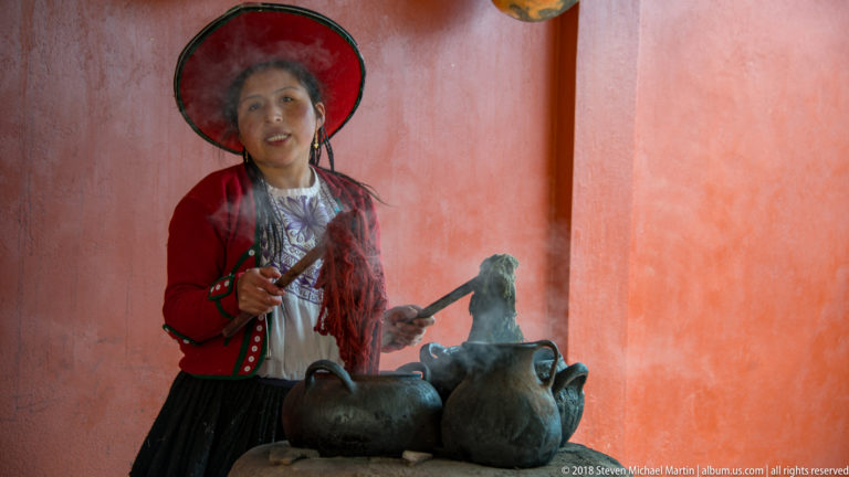 Woman in traditional highland clothing beside dark handled vessels emitting steam or smoke during a textile craft demonstration.