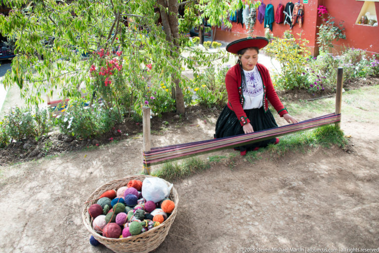 Woman in traditional highland clothing behind stretched warp threads, with a basket of brightly colored yarn balls in front.