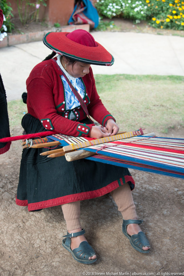 Andean woman in a broad red hat weaving a long multicolored textile outdoors with wooden rods controlling the threads.