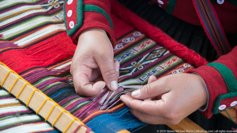 Weaver’s hands separating fine multicolored threads beside a wooden weaving element and a patterned red textile.
