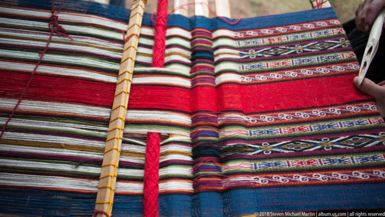 Red-dominant woven textiles with multicolored stripes and narrow geometric bands, crossed by a slender wooden rod.