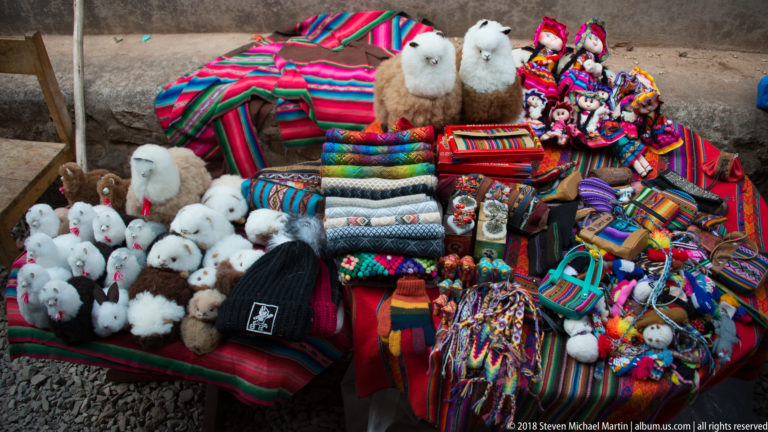 Market stall crowded with colorful woven goods, dolls, knitted accessories, and white and brown fluffy camelid souvenirs.