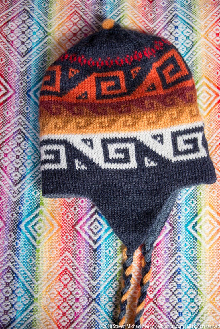 Knitted earflap hat with black, red, orange, brown, and cream geometric bands and braided ties on colorful woven cloth.