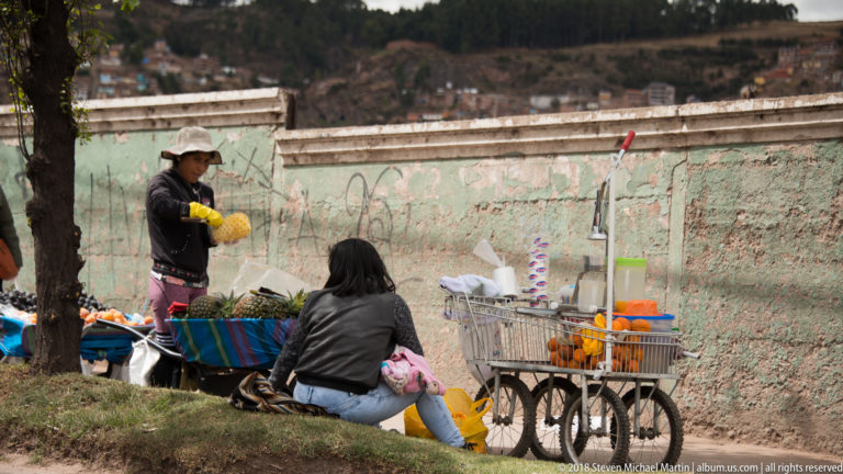 Two women selling fruit and produce beside a striped table covering, bags, and metal handcart near a weathered wall.