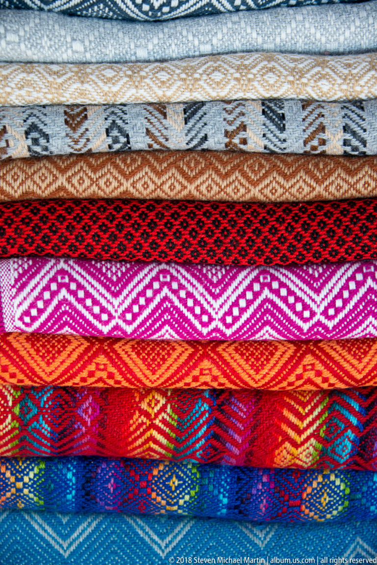 Stacked woven textiles with diamond and geometric bands in cream, gray, red, magenta, orange, blue, and turquoise.