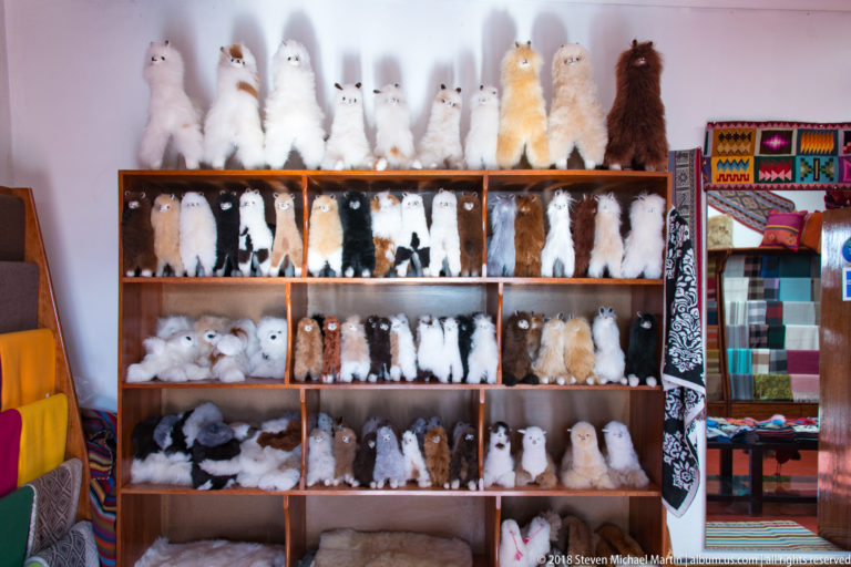 Wooden shelves filled with small fluffy camelid souvenir figures in white, brown, gray, and black beside colorful textiles.