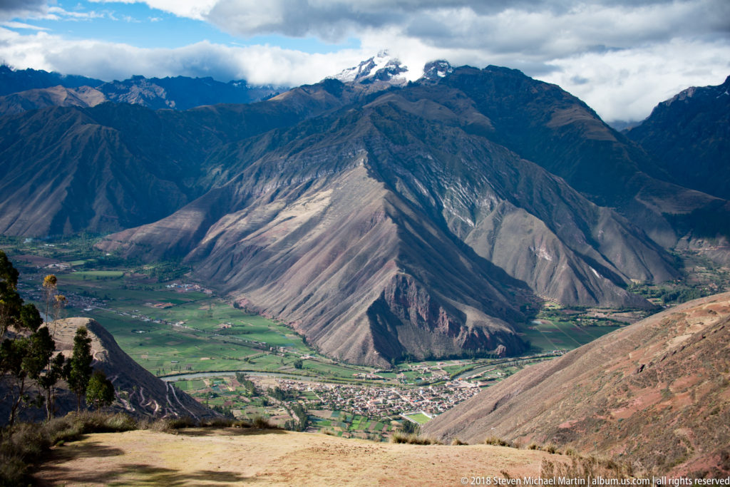 Broad Andean valley with pale eroded slopes, green fields, a town below, and darker mountains under clouds.