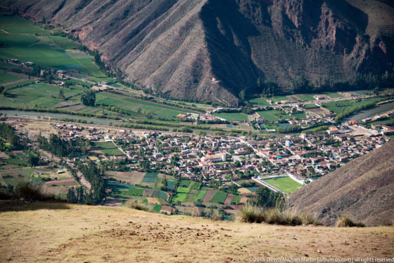 High-angle view of a compact Andean town, green patchwork fields, roads, and steep mountains enclosing the valley.