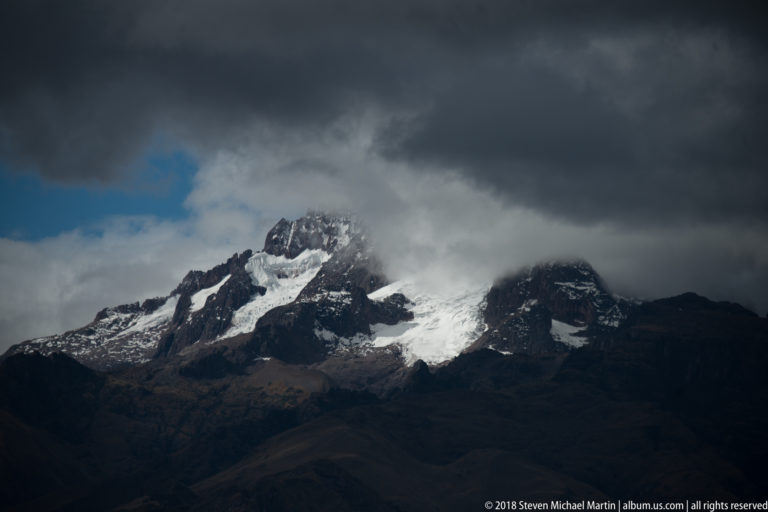 Rugged dark Andean mountain with snow patches beneath heavy gray storm clouds and a small opening of blue sky.