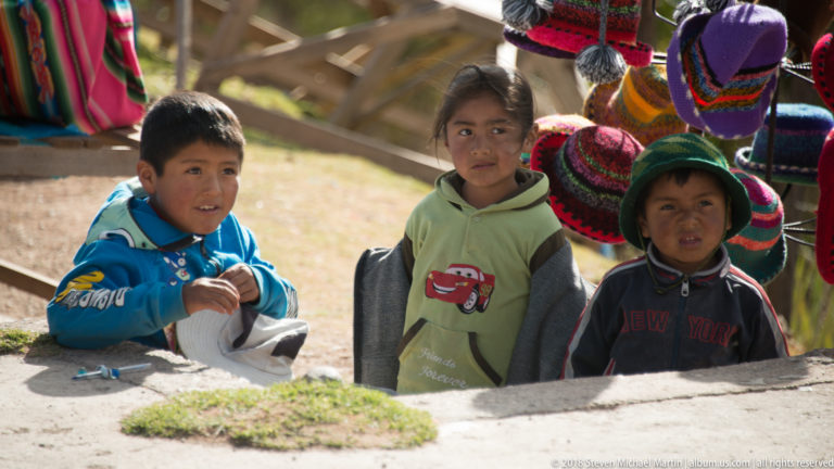 Three children in layered clothing stand beside brightly colored knitted hats at an Andean craft-market setting.