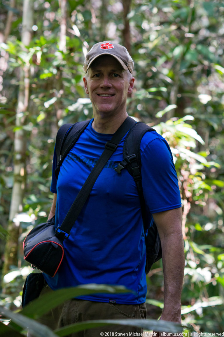 Smiling visitor in blue shirt and cap wearing a backpack and camera bag in rainforest.