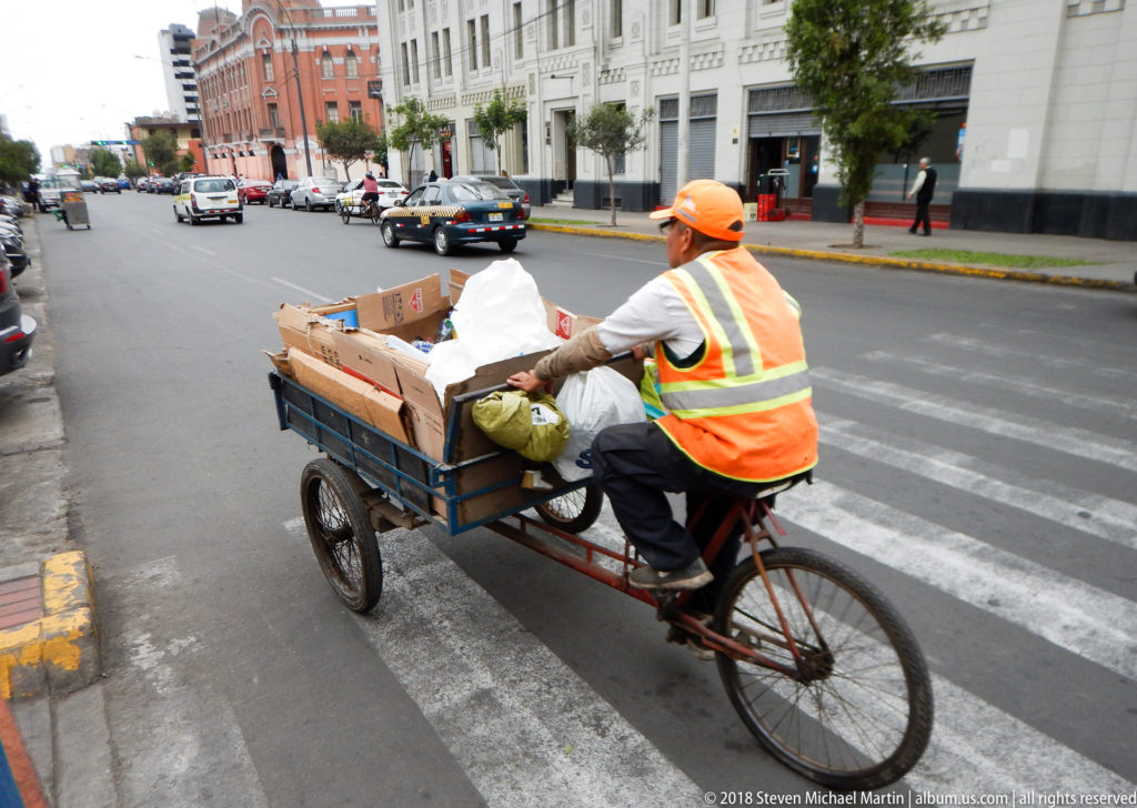 Worker in orange safety vest pedals a cargo tricycle loaded with boxes and bags across a Lima intersection.
