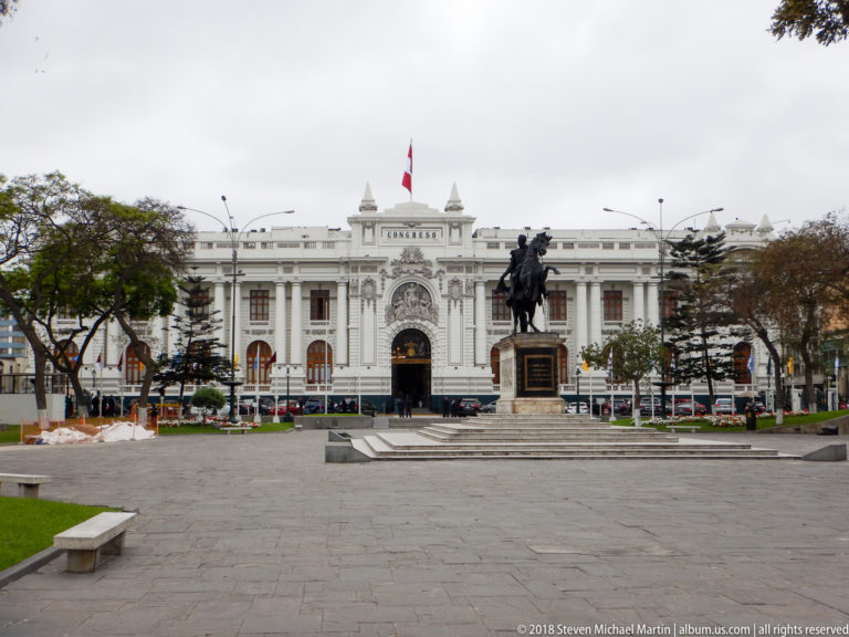 White Municipal Palace of Lima across a plaza, with Peruvian flag and an equestrian monument in the foreground.