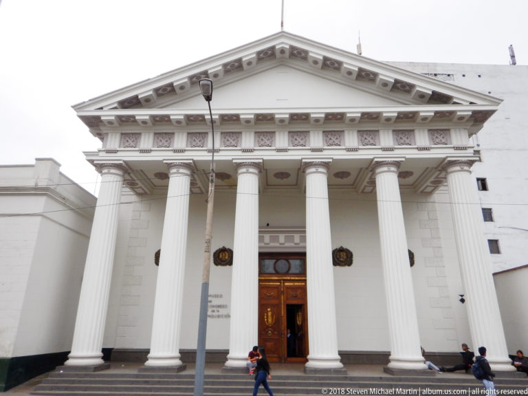 White neoclassical façade with four tall fluted columns, triangular pediment, decorative frieze, and central wooden doorway.
