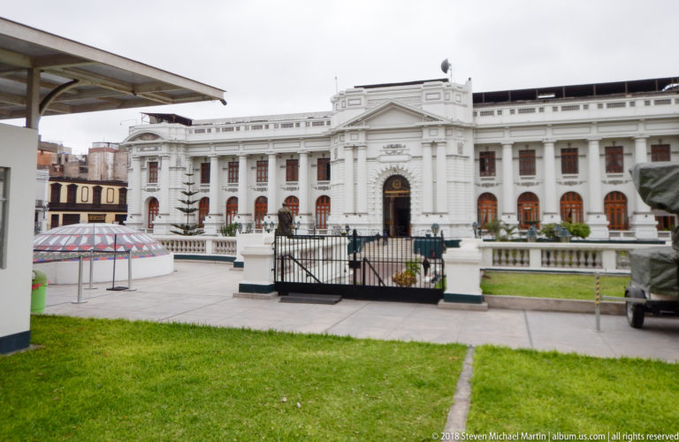 Large white neoclassical public building behind a gated lawn and forecourt, with repeated windows and a central pedimented entrance.