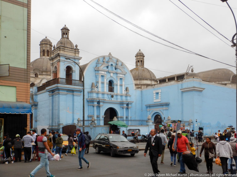 Blue historic church complex with domes and tower elements beside a crowded Lima street of pedestrians, vendors, and cars.