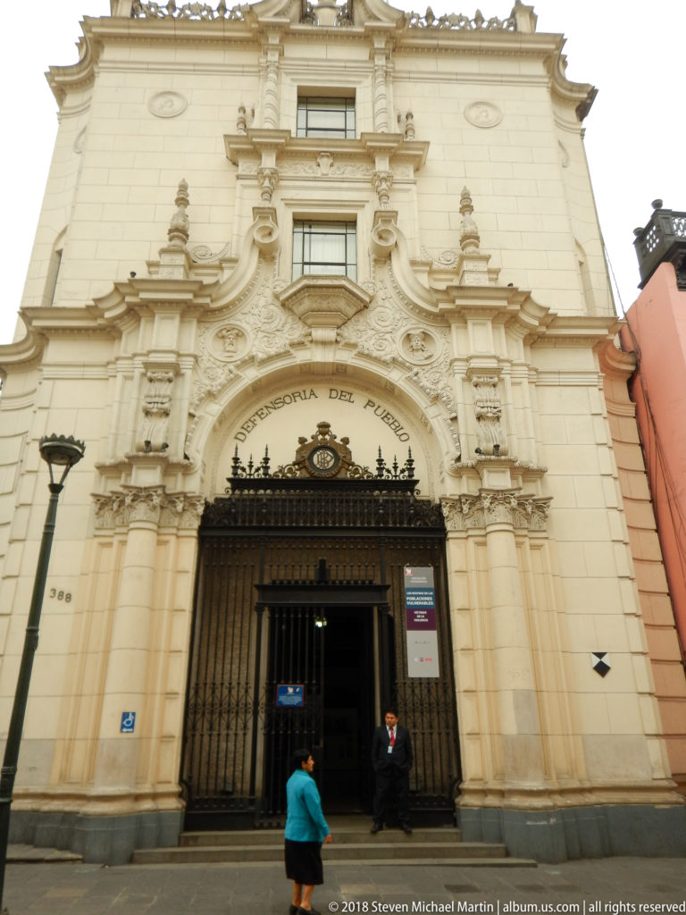 Pale ornate church façade with sculpted scrollwork and niches above a black entrance gate, with a person standing below.