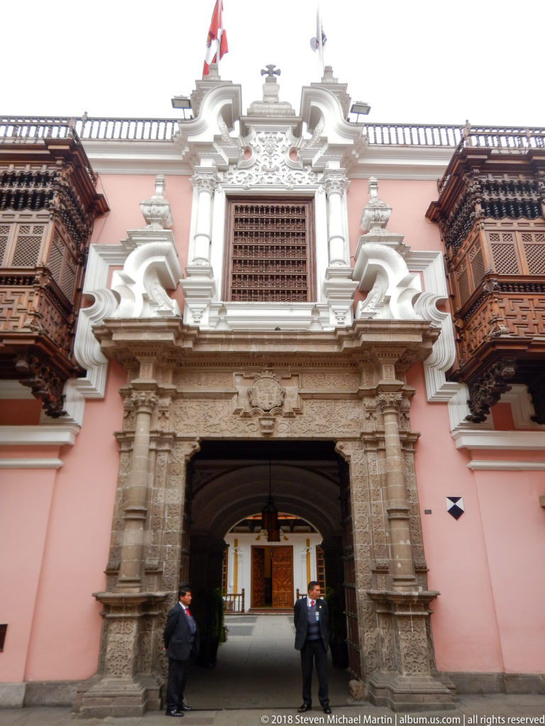 Front view of Palacio de Torre Tagle stone entrance with carved wooden balconies, pink façade, and two guards at street level.
