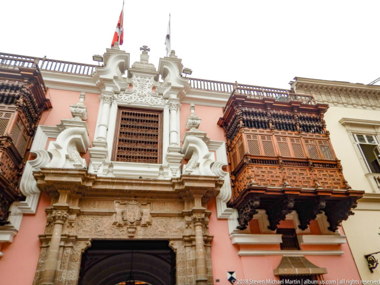 Low-angle view of Palacio de Torre Tagle with ornate stone portal, pink walls, carved enclosed wooden balconies, and Peruvian flag.