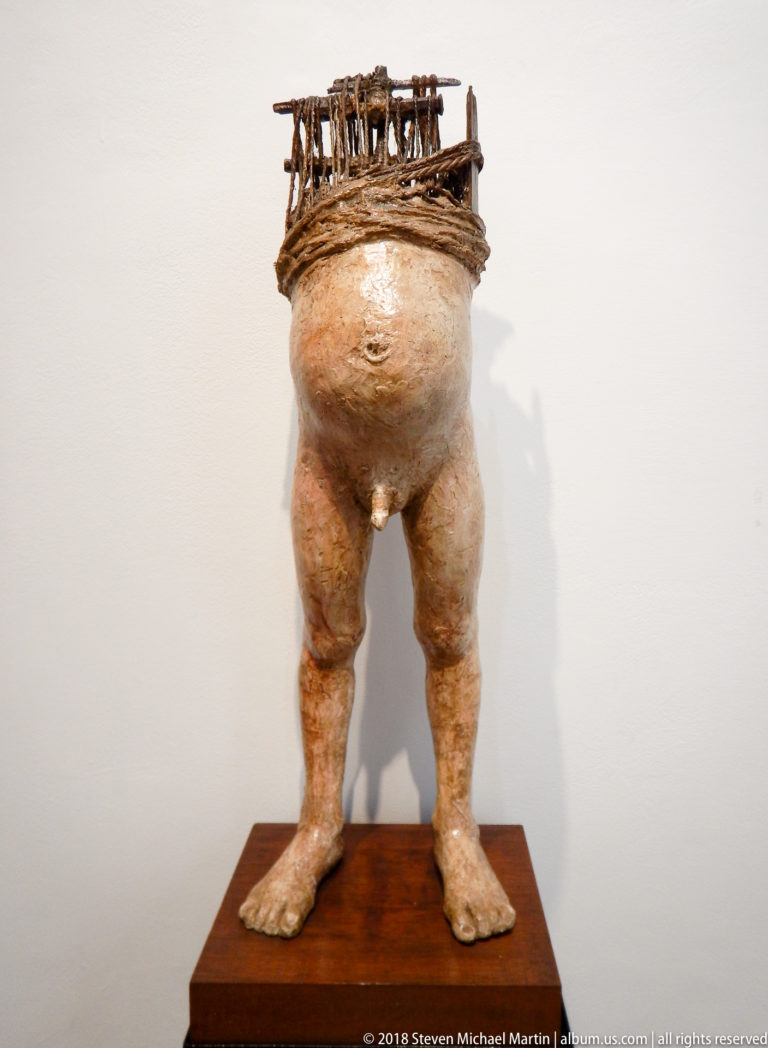 Roughly modeled standing human torso and legs topped by a small rack-like structure, displayed on a wooden pedestal.