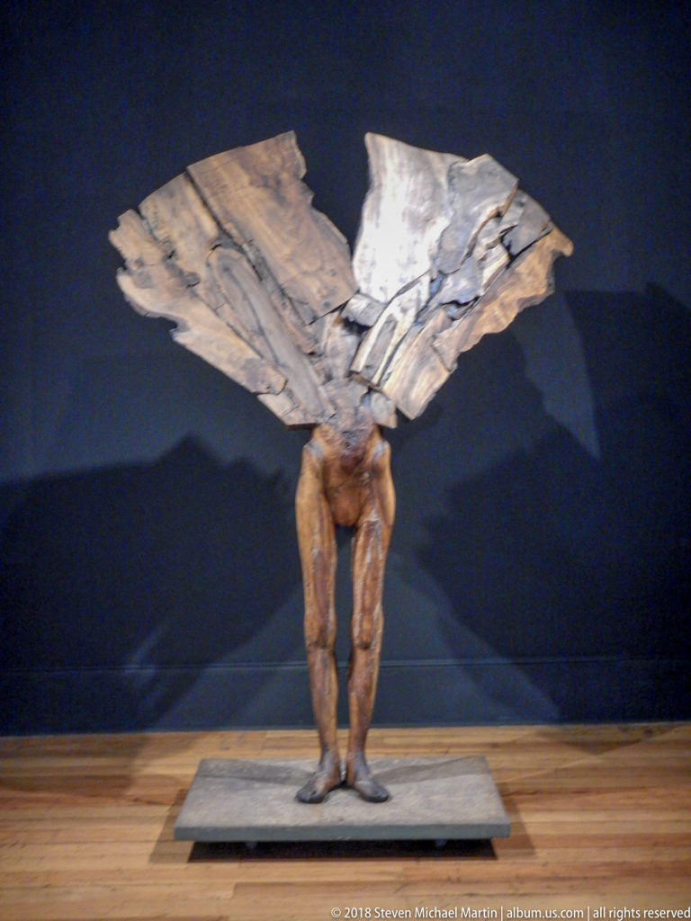 Standing sculpted figure bends forward beneath a broad pale folded form, displayed on a low platform against a dark wall.