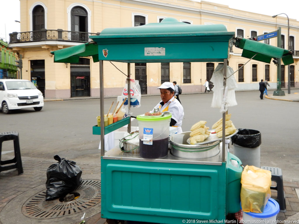 Vendor working at a green-canopied street food cart with containers and corn beside a central Lima street.