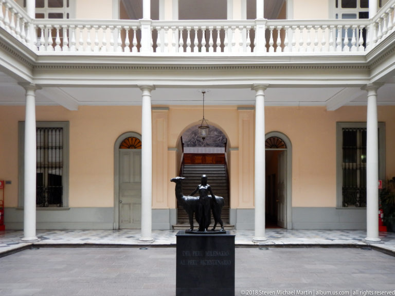 Bronze equestrian statue centered in a pale courtyard with columns, upper balustrades, doorways, and a staircase behind it.