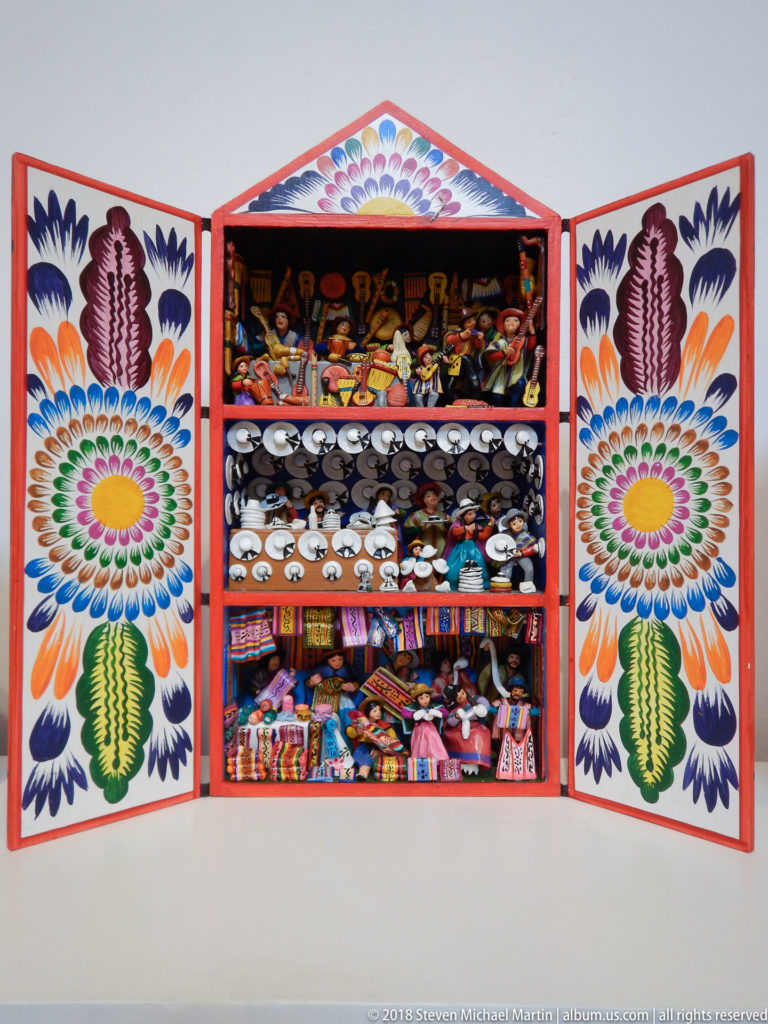 Three-tier open retablo with rows of pale miniature masks in the center and assorted figures and goods above and below.