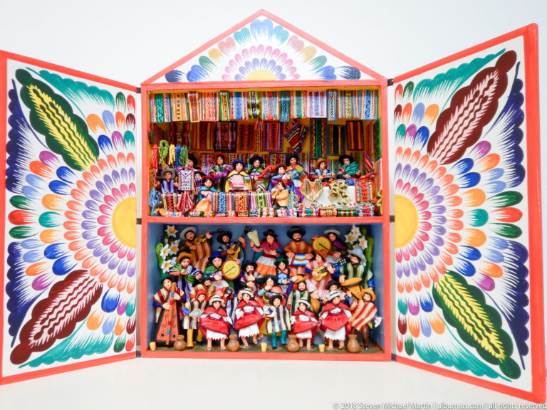Open floral-painted retablo with textile vendors on the upper tier and a crowded group of dancers and musicians below.