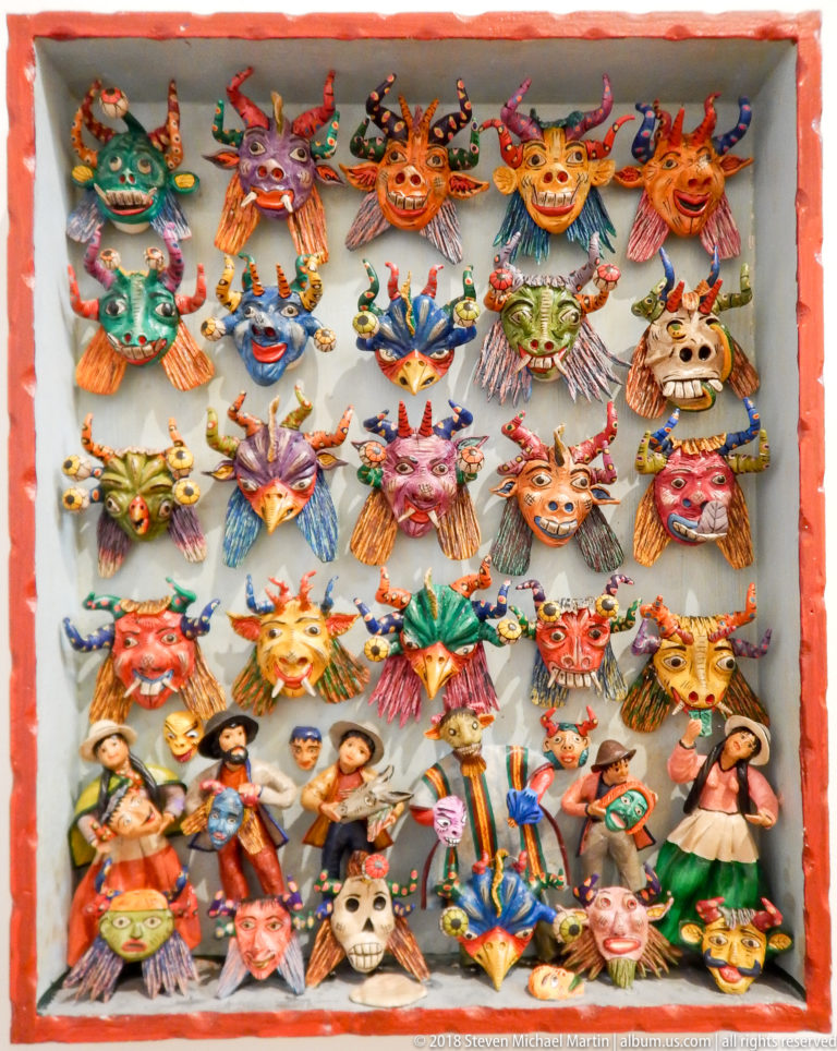 Framed display filled with rows of colorful miniature masks showing horned, human, animal, and grotesque faces.