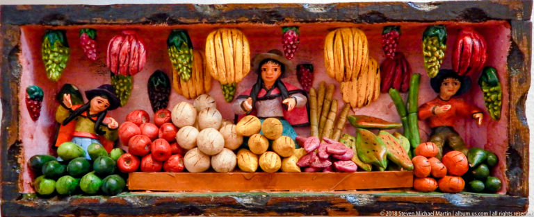 Relief market scene with three vendors behind piles of produce and multicolored ears of corn hanging overhead.