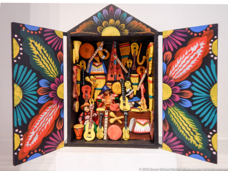 Small open retablo packed with miniature musical instruments and figures, bordered by black doors painted with bright leaves and flowers.