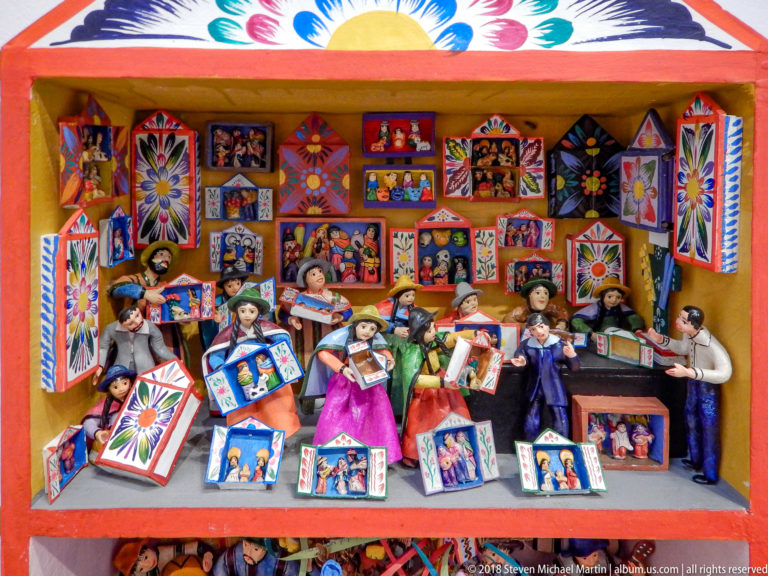 Colorful folk-art diorama with standing figures surrounded by many miniature framed pictures and decorated panels.