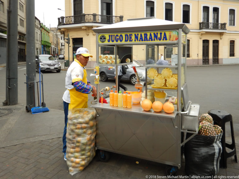 Vendor beside a stainless-steel orange-juice cart stocked with oranges and bottled juice on a Lima street.