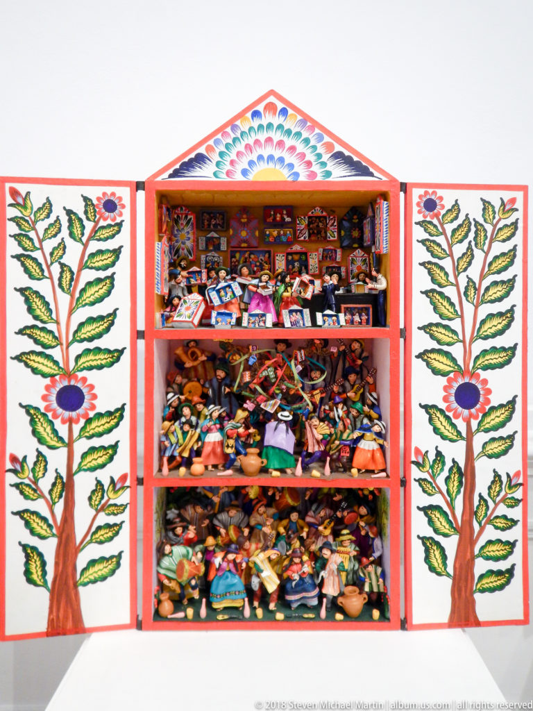 Open retablo with three crowded shelves of miniature goods and figures, framed by doors painted with flowering trees.