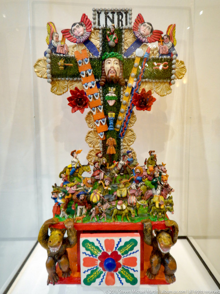 Colorful devotional crucifix with Christ, INRI plaque, small figures, flowers, birds, and densely decorated tiered base.