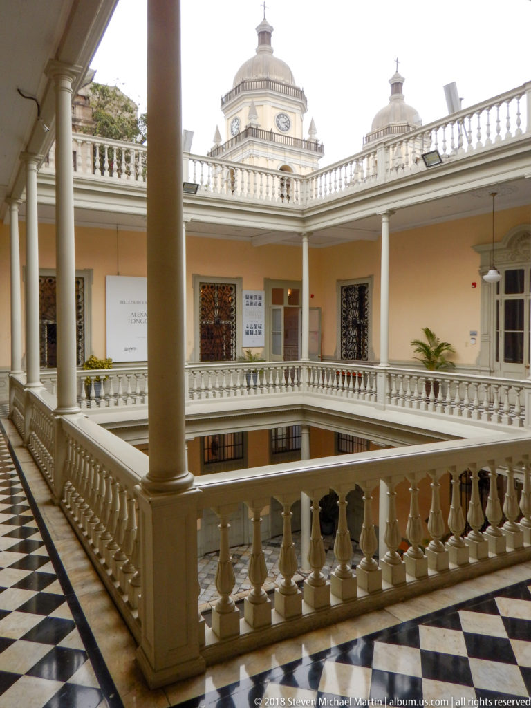 Two-story cream and white museum courtyard with columns, balustrades, black-and-white tile floor, and roof lantern beyond.
