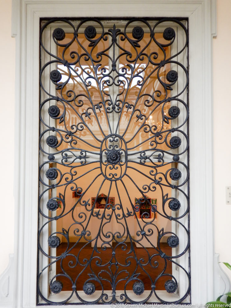 Black wrought-iron grille with central rosette and radiating fan-shaped scrollwork against a warm ocher background.