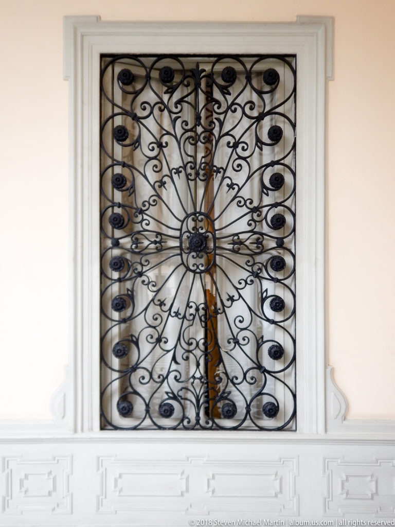 Black wrought-iron window grille with central rosette and symmetrical curling scrollwork on a pale wall.