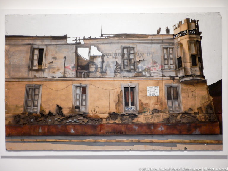 Wall-mounted artwork of a decaying two-story façade with graffiti, shuttered windows, peeling walls, and projecting balcony.