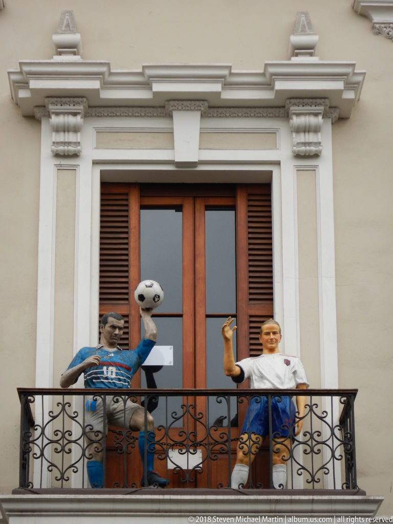 Football-themed decorative figures with a soccer ball posed on a wrought-iron balcony of a historic building.