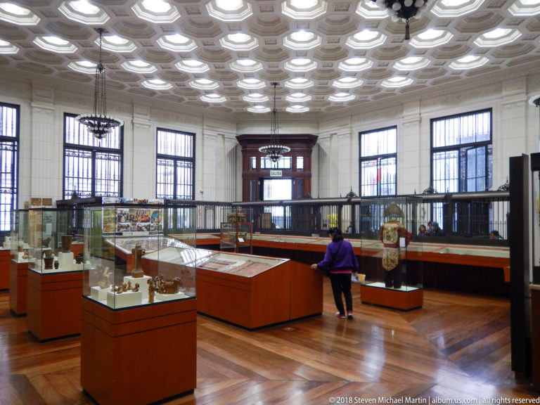 Bright museum gallery with glass artifact cases, polished wood floor, tall windows, balustrade, coffered ceiling, and one visitor.